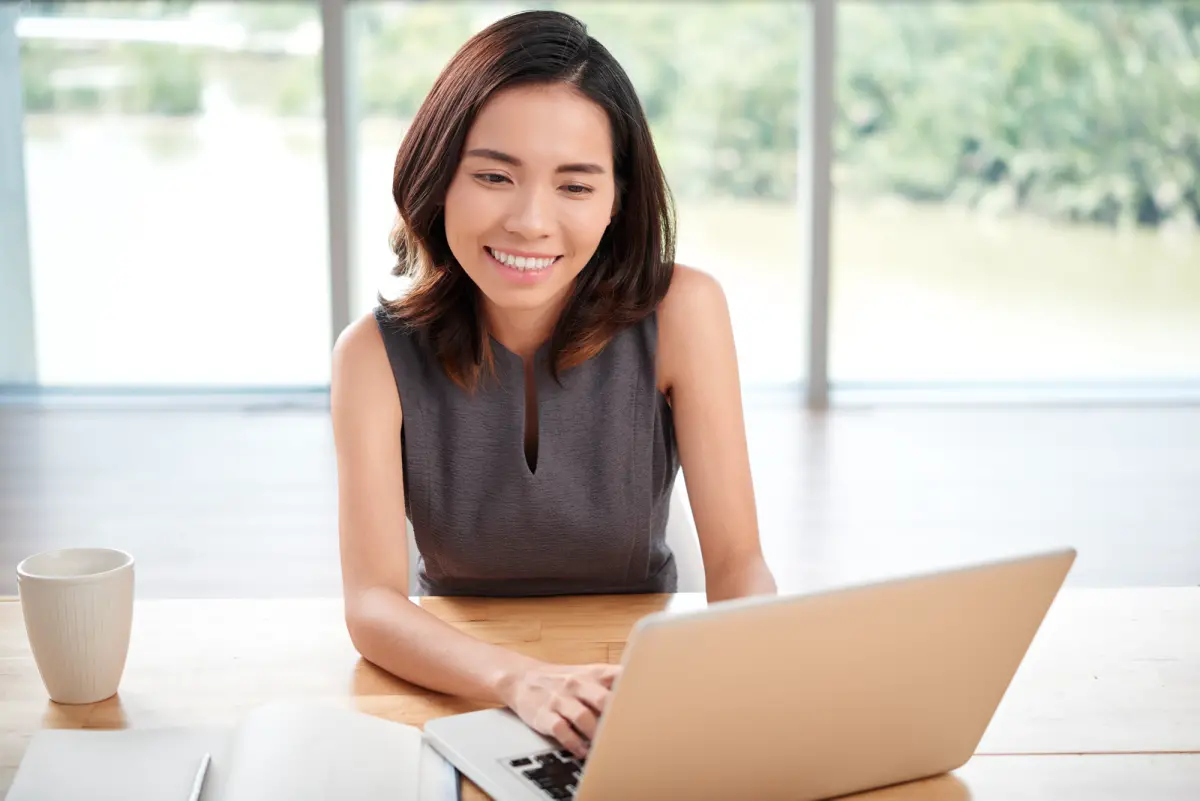 jeune femme brune souriant devant son ordinateur portable et tasse à café sur son bureau